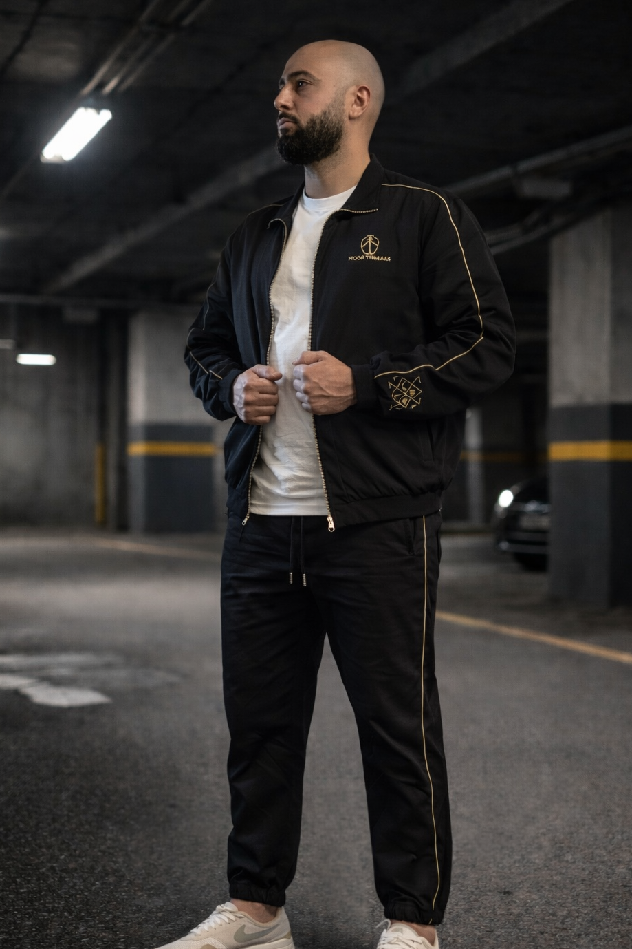 Man wearing a black jacket with gold accents in an underground parking garage.